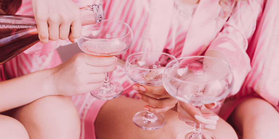 A close-up of champagne being poured into coupe glasses held by women in pink robes, suggesting a celebratory bridal party or bachelorette gathering at Bohemia Manor Farm Winery Resort.
