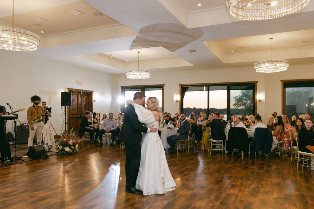 A bride and groom share their first dance in Bohemia Manor Farm's elegant ballroom reception space, with a live band performing to the left, guests seated at gold chiavari-chaired tables, crystal ring chandeliers glowing overhead, and a warm sunset visible through the ballroom's large picture windows.