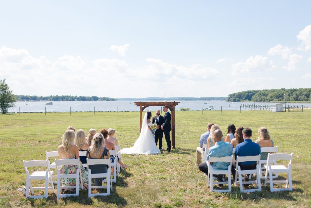 A couple exchanges vows beneath a rustic wooden arbor at Bohemia Manor Farm's waterfront ceremony lawn, with guests seated in white folding chairs on a sun-drenched green lawn, a broad river, wooded shoreline, and private dock stretching out behind them under a bright summer sky.