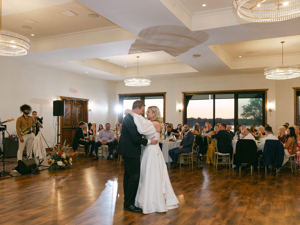 A bride and groom share their first dance in Bohemia Manor Farm's ballroom, with a live band, seated guests, crystal chandeliers, and a golden sunset waterfront view through the windows behind them.
