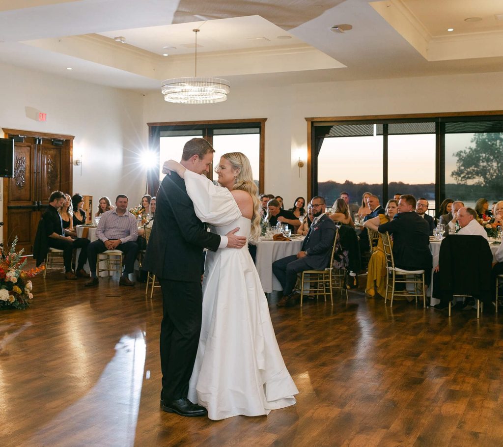 A bride and groom share their first dance in Bohemia Manor Farm's ballroom, with a live band, seated guests, crystal chandeliers, and a golden sunset waterfront view through the windows behind them.