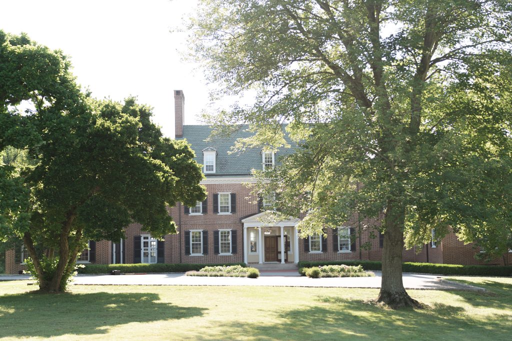 Front exterior of the historic brick manor house at Bohemia Manor Farm in Chesapeake City, Maryland, framed by towering mature trees, with black shutters, a patinated copper roof, columned entry portico, and sweeping green lawns.