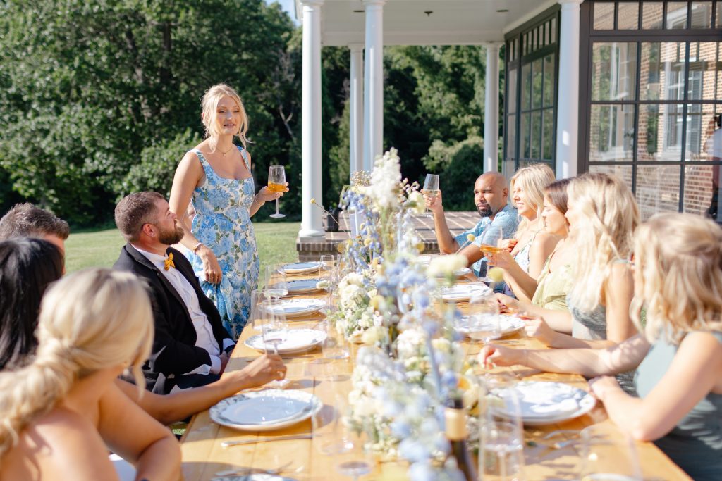 A woman in a blue floral dress raises a toast at a sunlit outdoor long-table brunch on the columned porch at Bohemia Manor Farm, surrounded by smiling guests seated along a flower-lined table.