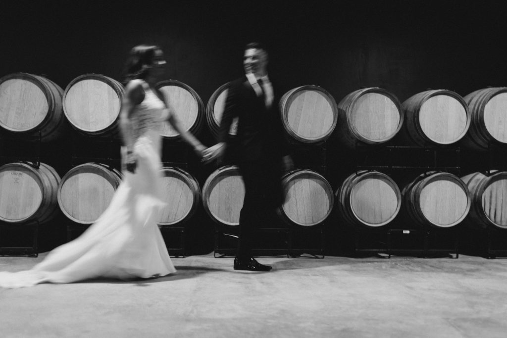 A black and white motion-blur photograph of a bride and groom dancing before stacked oak wine barrels in Bohemia Manor Farm's barrel room — one of the venue's distinctive and atmospheric wedding reception spaces.
