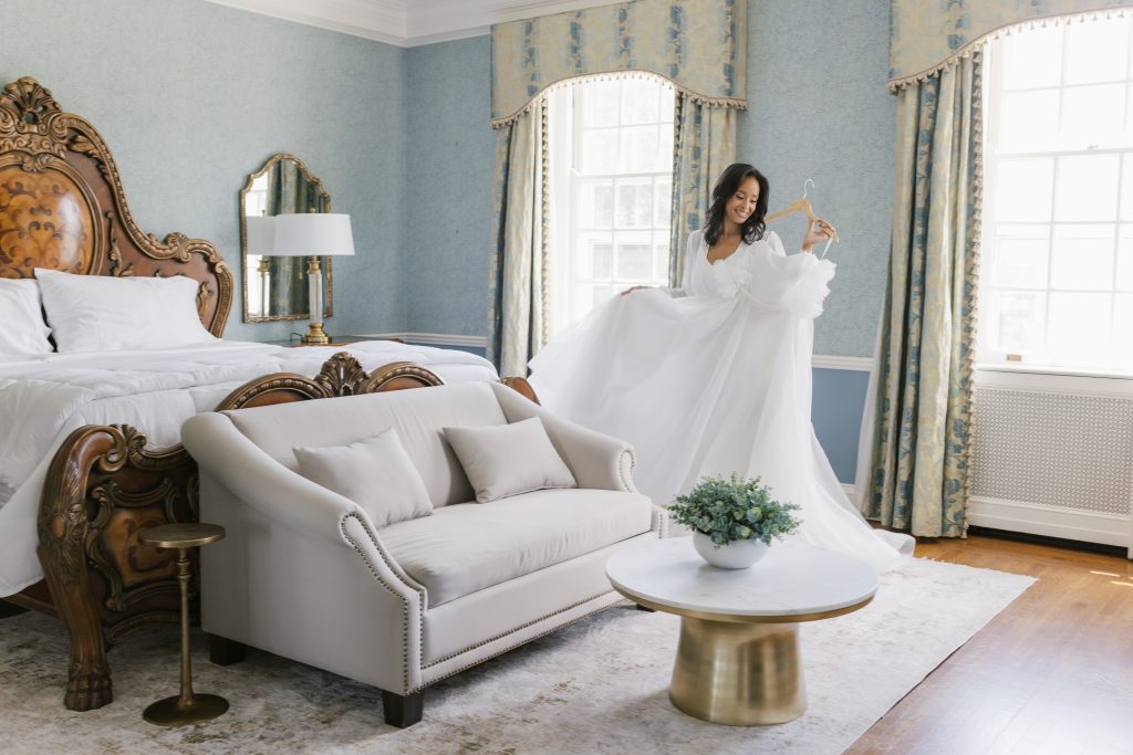 A bride smiles as she holds up her flowing white wedding gown in an elegantly appointed guest room at Bohemia Manor Farm, featuring powder blue walls, ornate carved wood furniture, gold accents, and floral drapes.