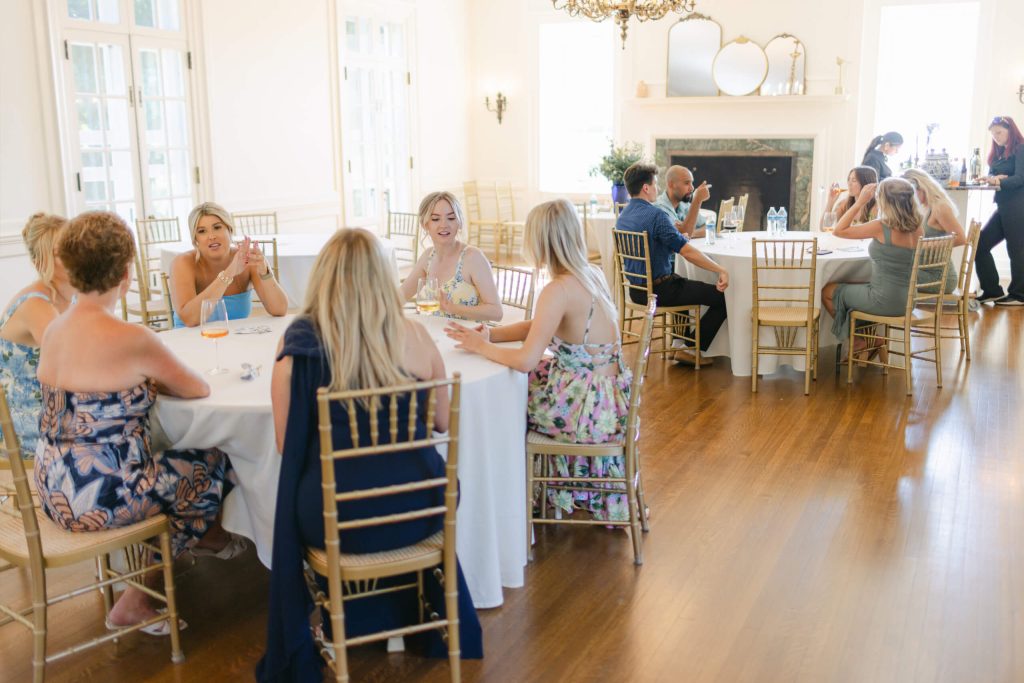 Guests socializing at round tables with gold chiavari chairs in Bohemia Manor Farm's sunlit drawing room, with the green marble fireplace and antique gold mirrors on the mantel visible behind them.