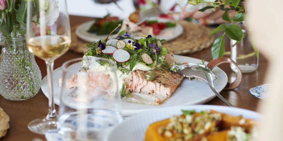 A whole roasted salmon fillet garnished with edible flowers, fresh herbs, sliced radishes, and cream sauce on a white platter, surrounded by a glass of white wine, crystal vases with greenery, and additional dishes on a rustic wooden dining table, representing farm-to-table cuisine at Harvest & Tide at Bohemia Manor Farm.