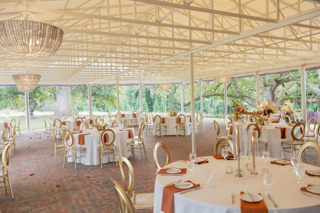 A sunlit open-air reception pavilion at Bohemia Manor Farm, elegantly set with round white-draped tables, gold oval-back chairs, burnt orange napkins, and autumn floral centerpieces — crystal bead chandeliers hang from white exposed trusses above a herringbone brick floor, with sweeping views of mature trees and lush grounds through the open sides.