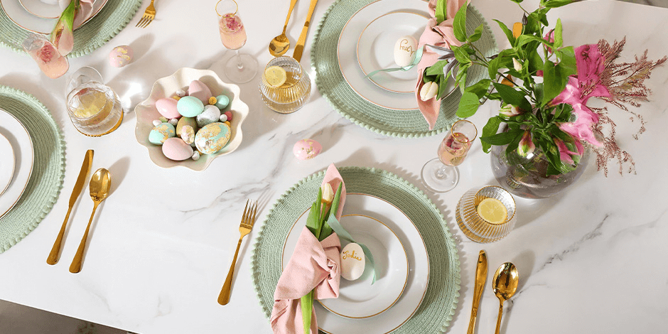 Overhead flat-lay of an elegantly styled Easter brunch table at Harvest & Tide at Bohemia Manor Farm — sage green woven chargers, gold-rimmed white china, gold flatware, and blush pink napkins tucked with fresh tulips and personalized painted egg place cards at each setting, a shell-shaped bowl of pastel Easter eggs as a centerpiece, crystal glassware with lemon garnishes, and a lush pink and green floral arrangement on a white marble table.