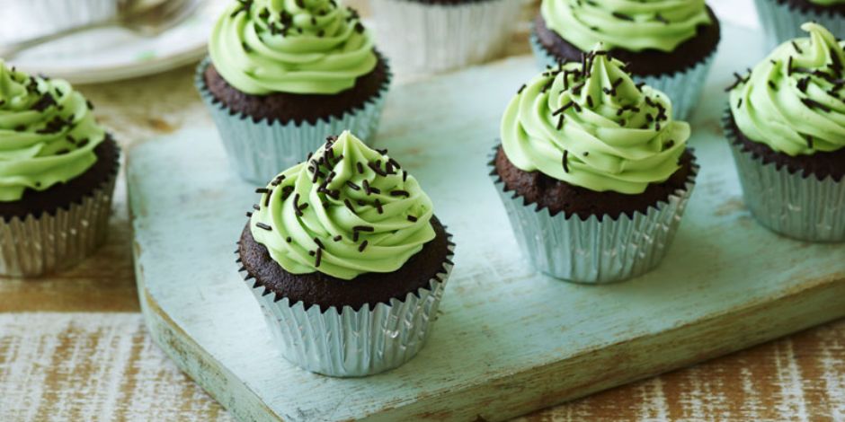 Chocolate cupcakes with swirled green buttercream frosting and dark chocolate sprinkles in silver foil liners, arranged on a mint green distressed wood cutting board
