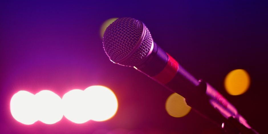 A close-up of a handheld microphone on a stand under dramatic purple and white stage lighting with warm amber bokeh in the background, representing live entertainment at Bohemia Manor Farm Winery Resort.