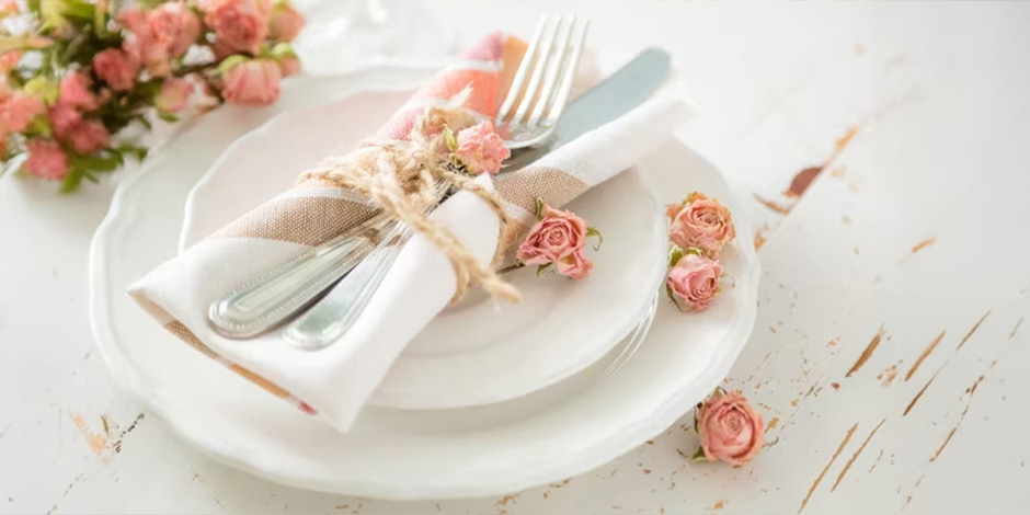 A table place setting with white plates, a linen napkin tied with twine and pink spray roses, and scattered rose buds on a white distressed wood surface.