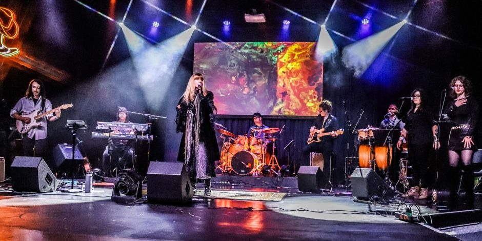 A full eight-piece band performs live on stage at Bohemia Manor Farm under dramatic beam lighting, with a female lead vocalist at center, a full band behind her, and a colorful abstract video display backdrop.
