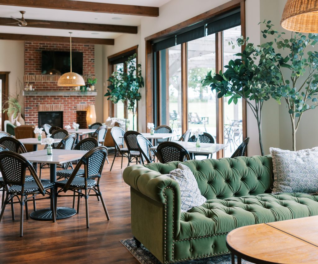 Bright, airy dining room at Bohemia Manor Farm with bistro-style tables, rattan pendant lighting, exposed wood beam ceiling, brick fireplace, large glass doors opening to an outdoor patio, and a tufted green velvet sofa in the foreground.