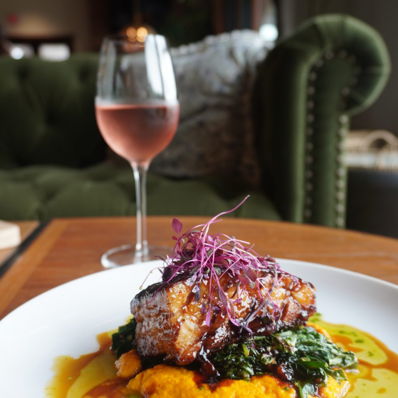 A plated farm-to-table entrée of glazed braised pork belly over butternut squash purée with sautéed greens and purple microgreens, paired with a glass of rosé wine — served at a wooden table in the intimate dining room of Harvest & Tide at Bohemia Manor Farm, the restaurant's signature green tufted velvet sofa softly visible in the background.