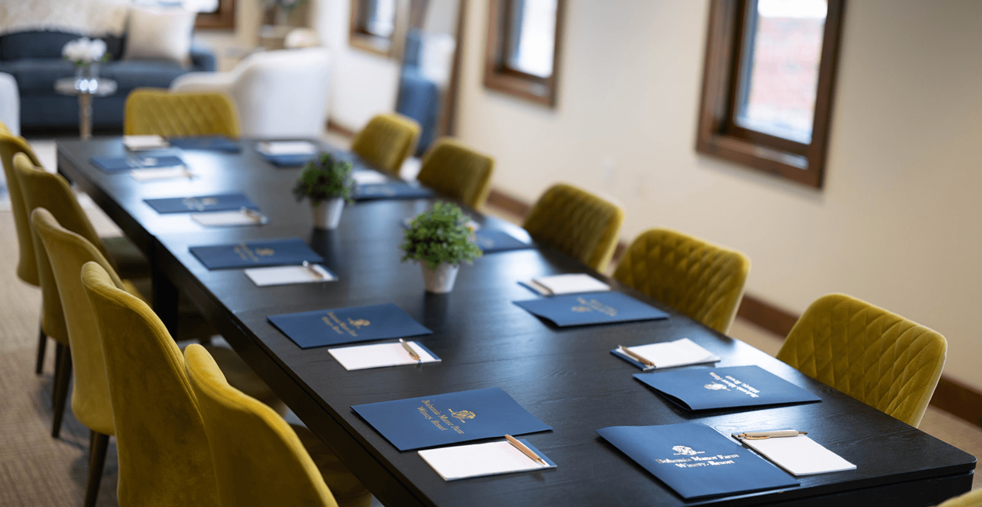 A corporate meeting table at Bohemia Manor Farm set with branded navy folders, notepads, and pens at each place, with yellow upholstered chairs and a potted plant centerpiece.