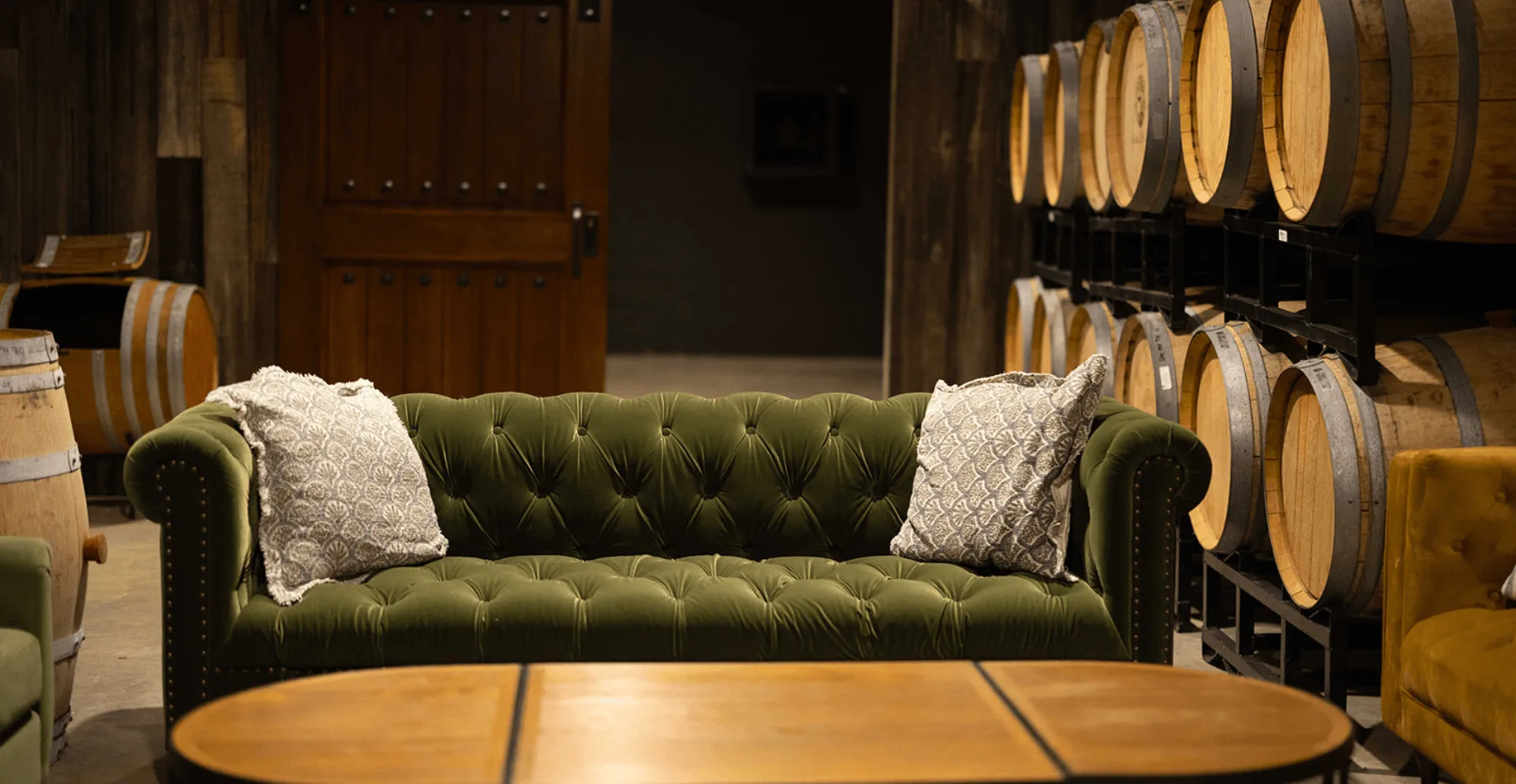 A forest green tufted velvet Chesterfield sofa with white throw pillows in Bohemia Manor Farm's barrel room, surrounded by stacked oak wine barrels and a rustic wooden door beyond.