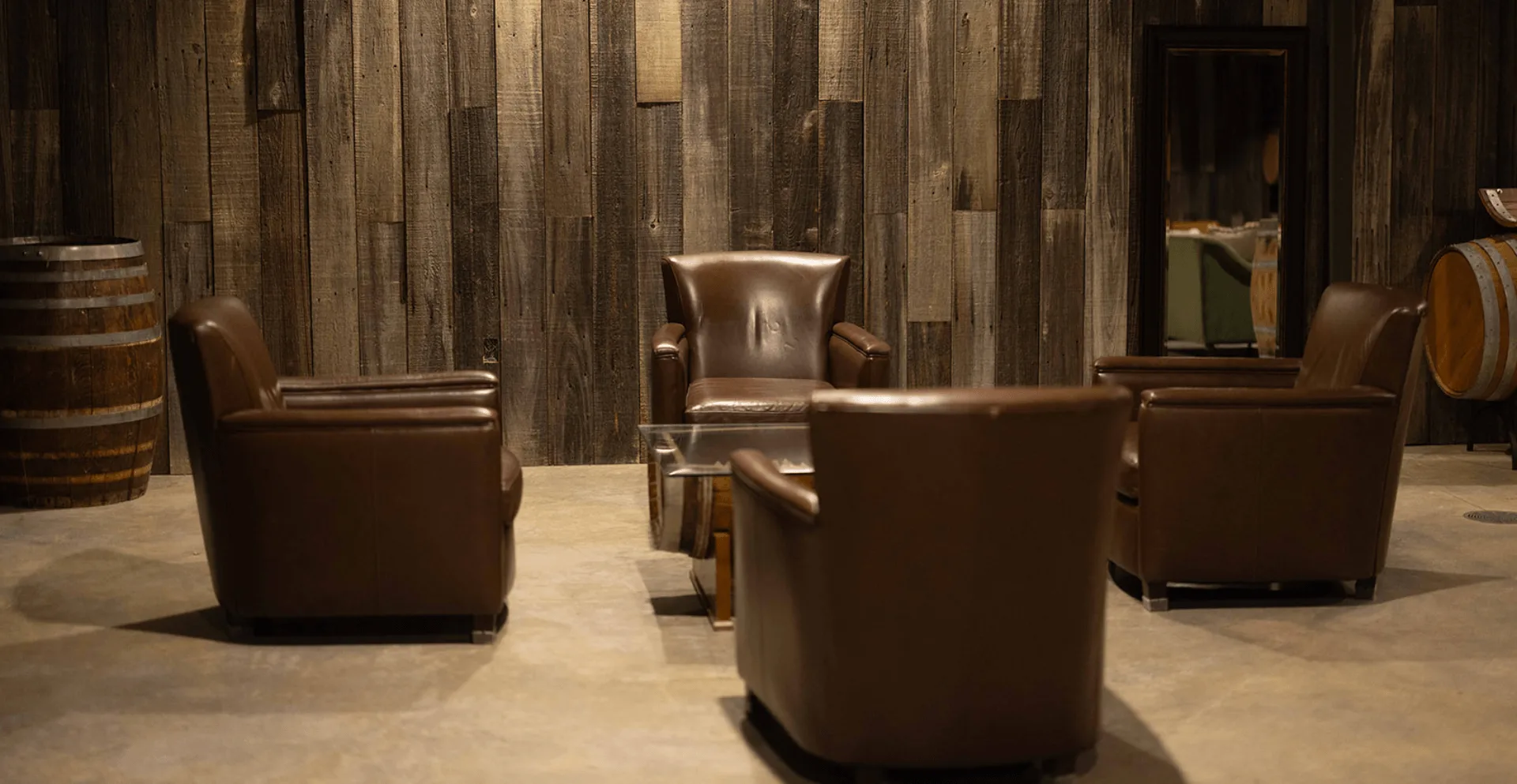 Four brown leather club chairs around a barrel-base glass coffee table in Bohemia Manor Farm's barrel room, with a reclaimed wood accent wall and oak wine barrels on either side.