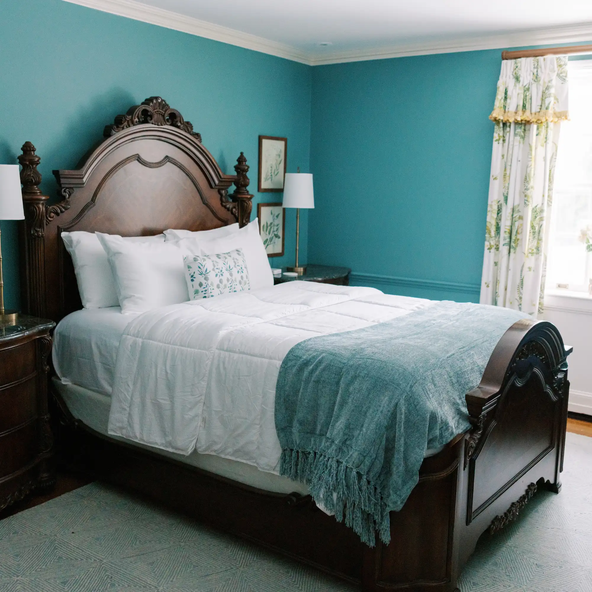 A Bohemia Manor Farm guest room with a grand carved mahogany bed, white linens, teal throw, vibrant teal walls, botanical prints, and green floral curtains framing a bright window