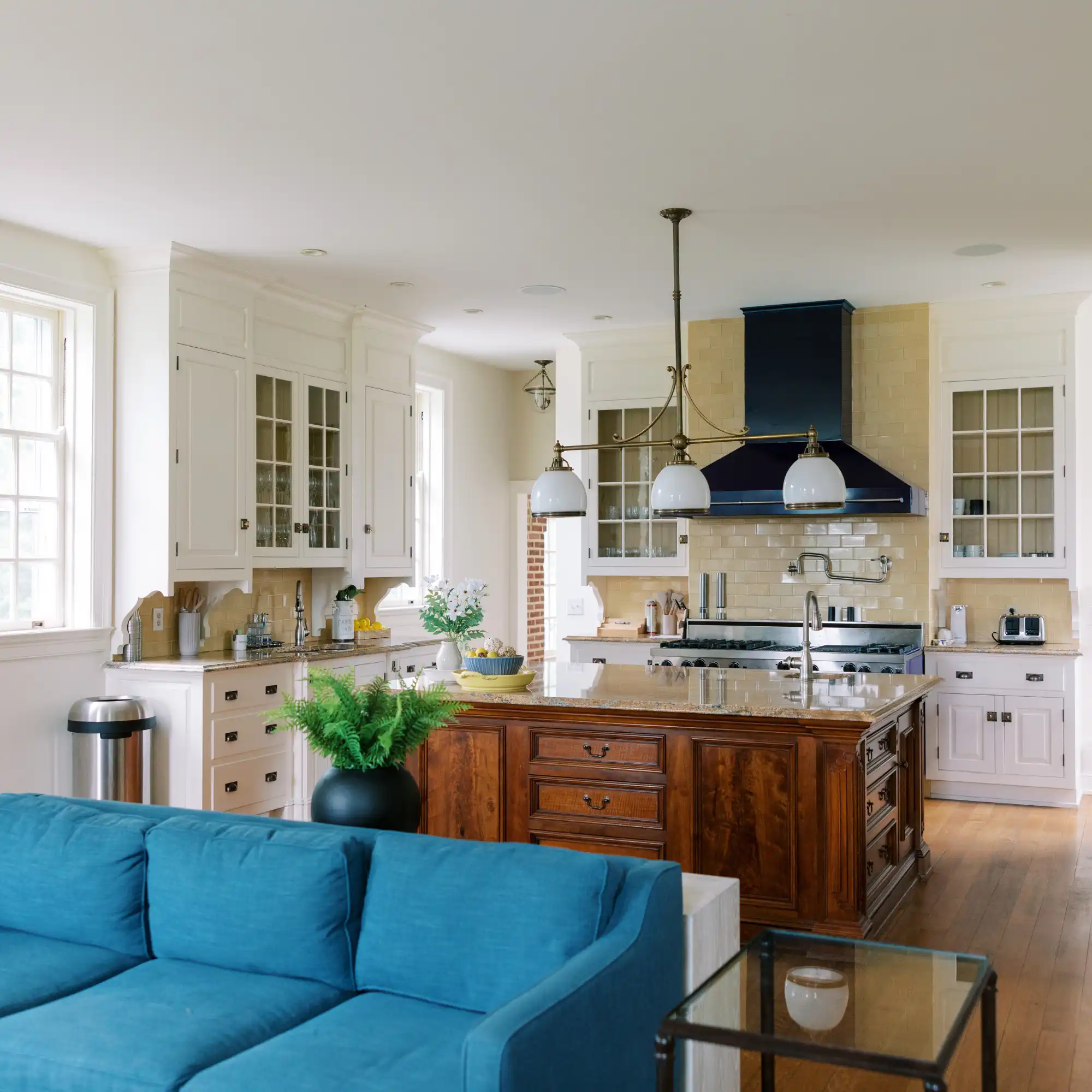Bohemia Manor Farm's manor kitchen with white glass-front cabinets, an antique walnut island with granite top, navy range hood, brass pendant lights, and a teal sofa in the adjoining open living area.