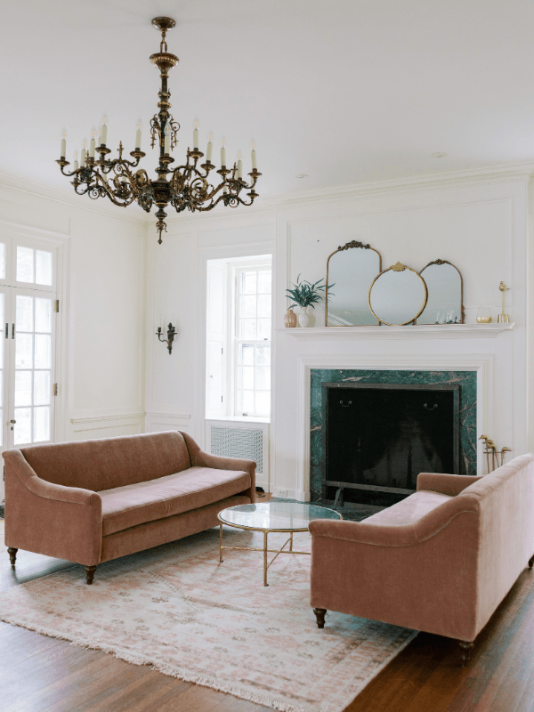 Bohemia Manor Farm's parlor with two dusty rose velvet sofas, a green marble fireplace, antique gold mirrors on the mantel, and a grand bronze candelabra chandelier above a blush rug on hardwood floors.