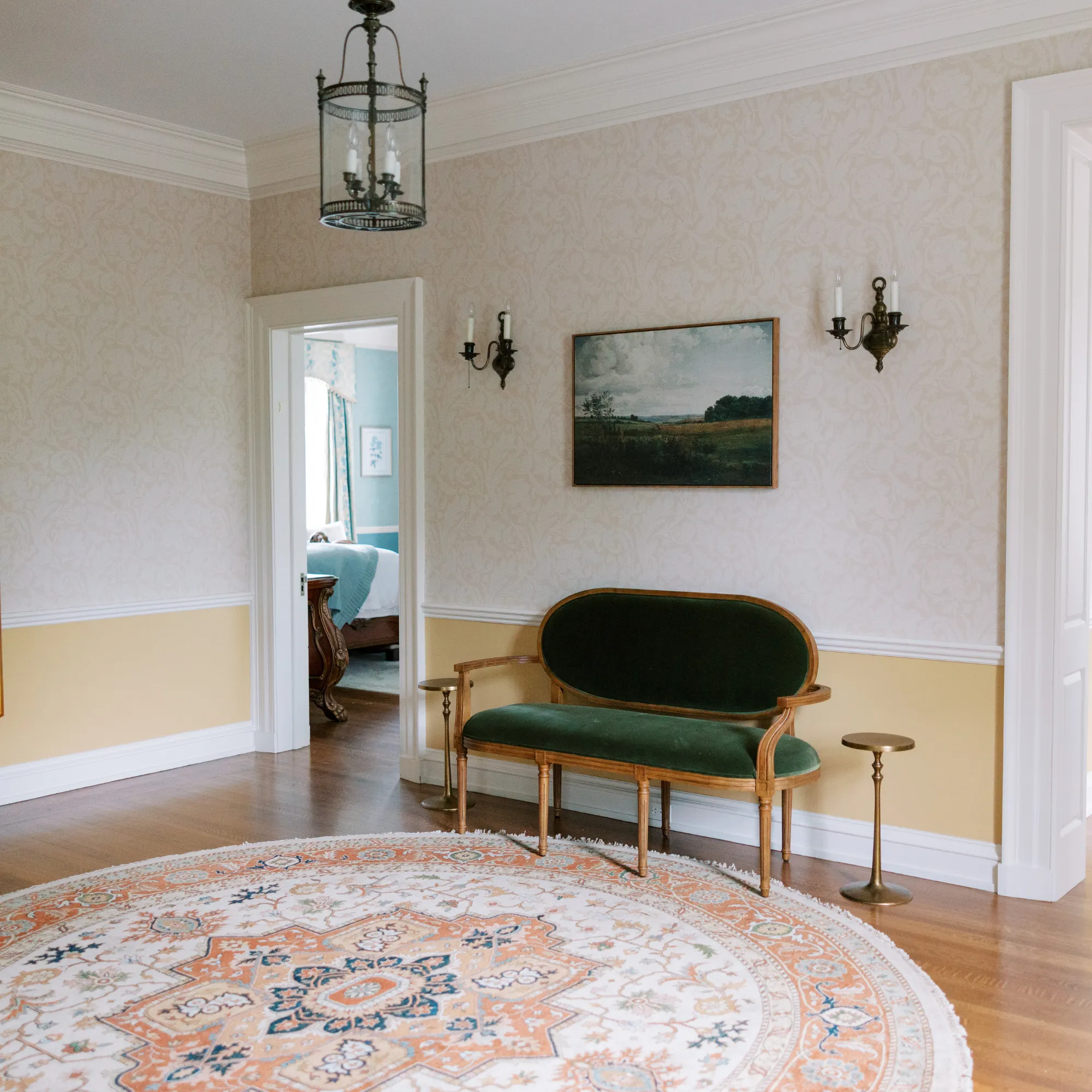 An elegantly appointed upper hallway in Bohemia Manor Farm's historic manor house, featuring cream damask wallpaper above warm golden yellow wainscoting, a green velvet Louis XVI-style settee with a gilded wood frame centered beneath a framed pastoral landscape painting and flanked by brass candle wall sconces — a round terracotta and cream Persian rug anchors the hardwood floor beneath a wrought iron lantern chandelier, with an open doorway offering a glimpse into a sunlit blue guest bedroom beyond. The open doorway is a lovely compositional detail worth noting — it invites the viewer deeper into the house and subtly communicates the scale and interconnectedness of the manor's interior spaces.
