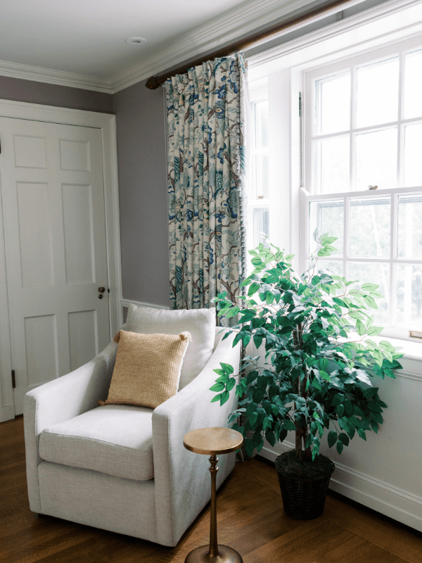 A guest room sitting corner at Bohemia Manor Farm with a cream armchair, brass side table, large ficus tree, and blue botanical print curtains framing a bright multi-pane window.