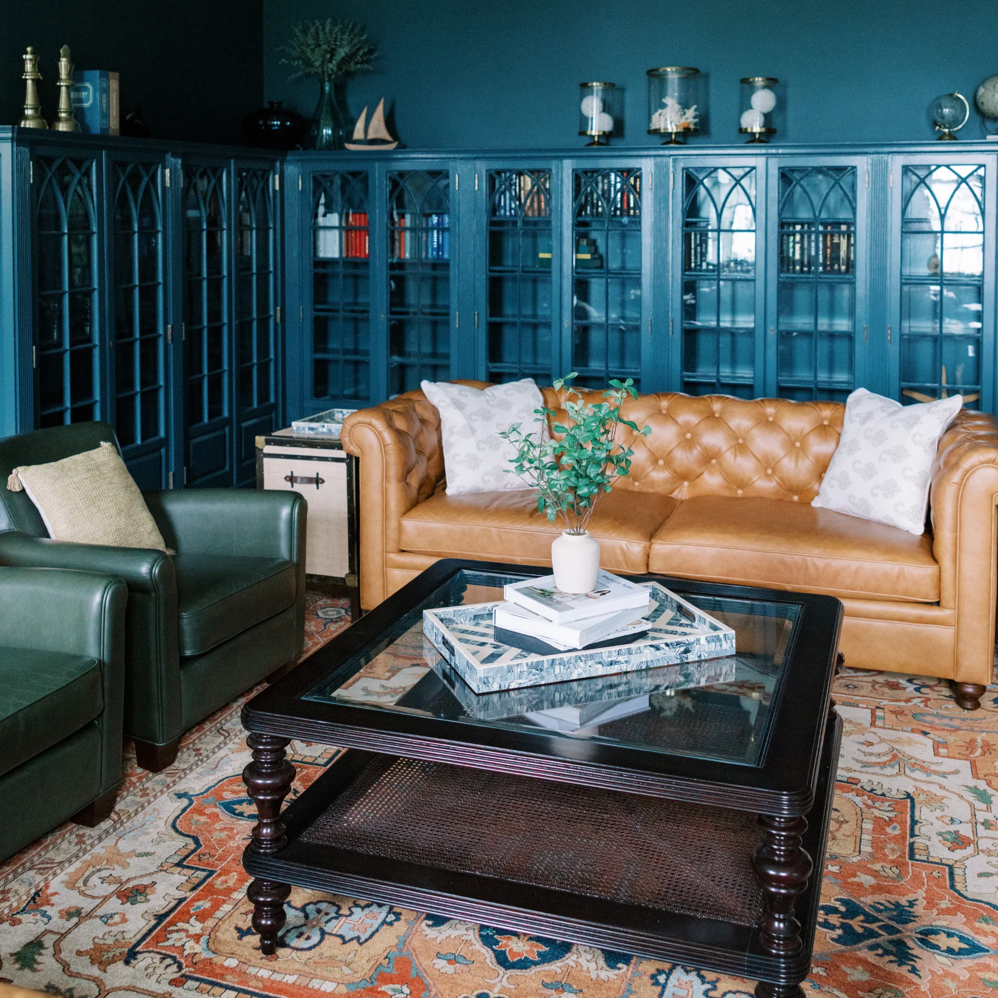 A common space at Bohemia Manor Farm, featuring a full wall of floor-to-ceiling teal-painted Gothic arch glass-front bookcases filled with books and curated maritime objects — flanking a tufted caramel leather Chesterfield sofa with white throw pillows and a deep green leather armchair, centered around a large black glass-top coffee table on a terracotta and blue Persian rug over hardwood floors.