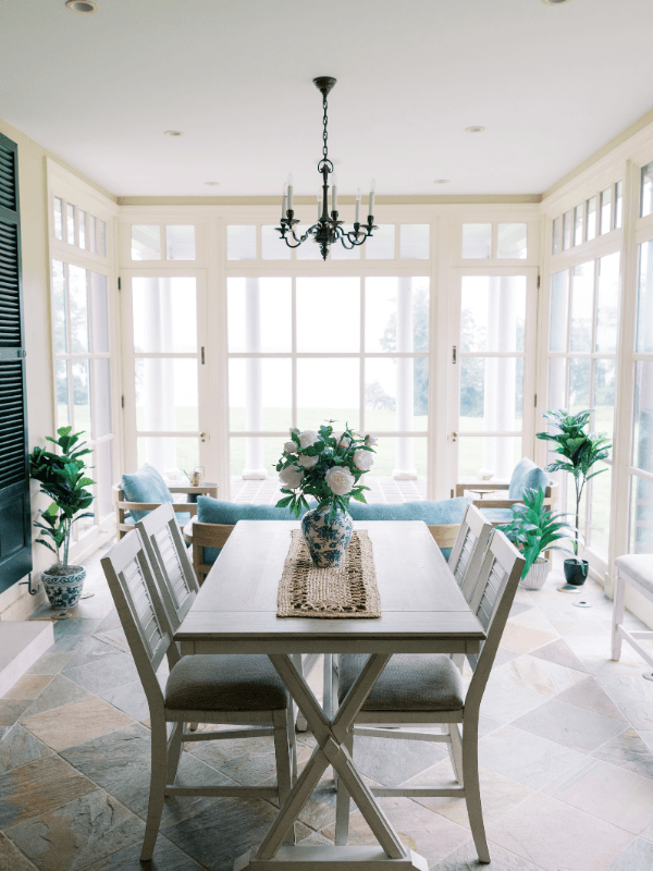 A sunlit sunroom dining space at Bohemia Manor Farm, centered around a white trestle table with a jute runner and blue and white chinoiserie vase of white roses, surrounded by gray upholstered chairs beneath a black iron candelabra chandelier — flanked by blue cushioned lounge seating, potted tropical plants, and a full wall of white-framed French doors and transom windows opening to the lush green grounds beyond.