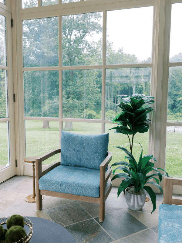 A serene sunroom corner at Bohemia Manor Farm, featuring a wood-framed lounge chair with blue patterned cushions and a tall tropical plant in a ribbed ceramic pot on slate tile flooring — floor-to-ceiling white-framed windows wrap around the space, framing an expansive view of lush green lawns and mature trees beyond.