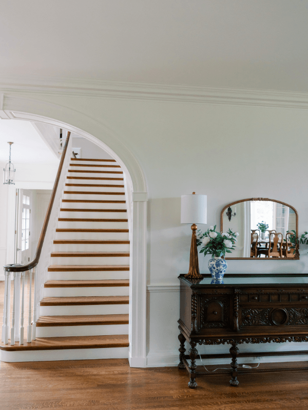 The elegant foyer of Bohemia Manor Farm's historic manor house, featuring a sweeping staircase with warm oak treads and white risers set within a graceful arched opening, alongside an ornately carved dark wood antique sideboard styled with a gold tassel table lamp, a blue and white chinoiserie vase filled with greenery, and a gilded arched mirror reflecting glimpses of the dining room beyond — all on rich hardwood floors beneath white crown molding.
