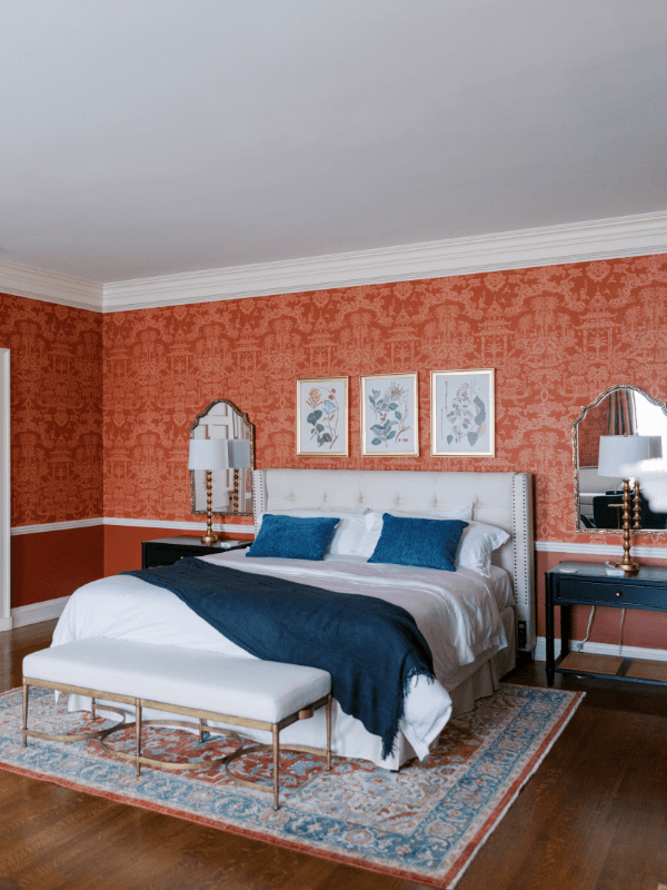 A beautifully appointed guest suite at Bohemia Manor Farm featuring bold red and cream chinoiserie-patterned wallpaper, a king bed with a cream tufted wingback headboard dressed in white linens and navy throw and pillows, matching dark navy nightstands with gold lamps and arched mirrors, three framed botanical prints centered above the bed, a white upholstered bench on a red and blue Persian rug, and warm hardwood floors throughout.