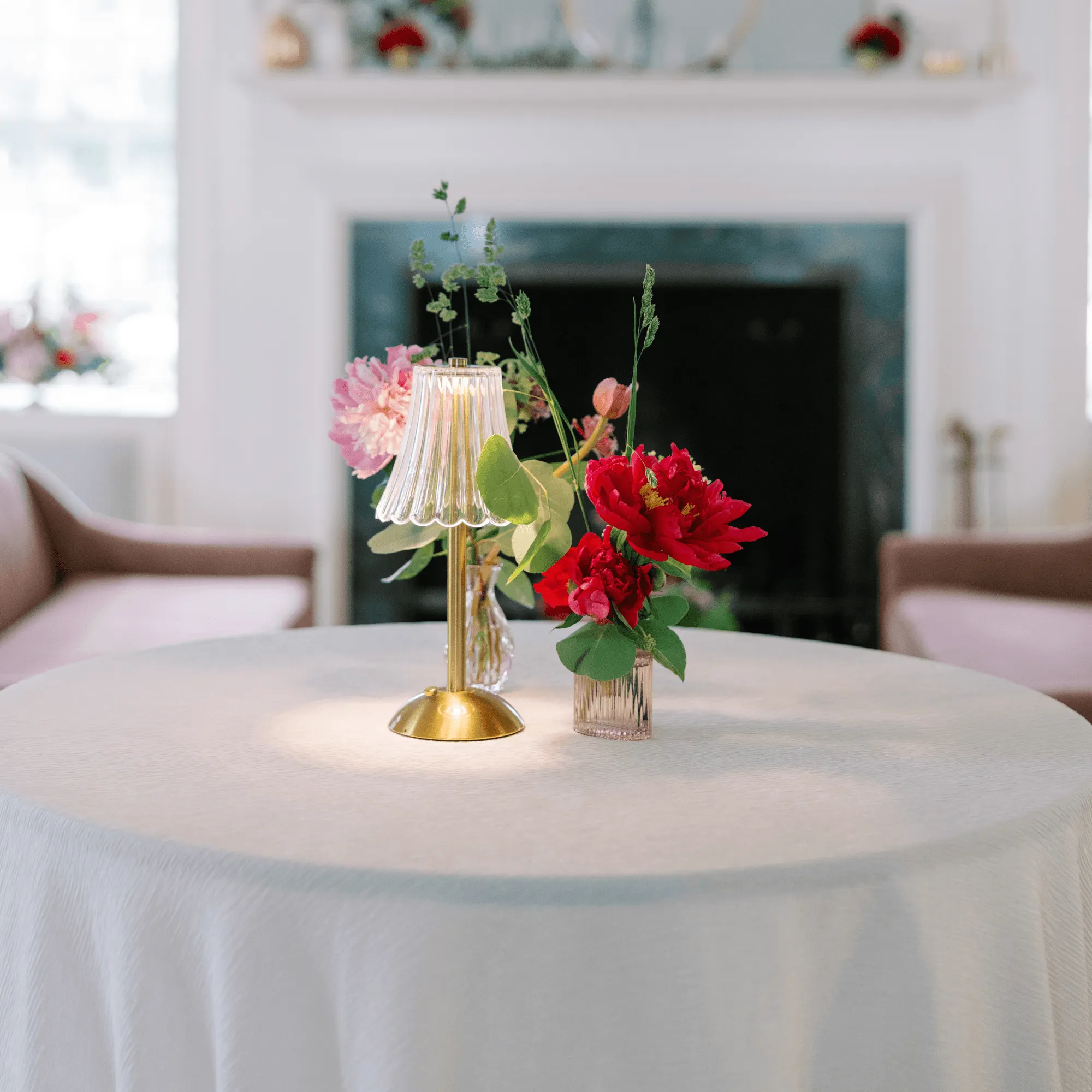 A close-up of a white linen-draped reception table at Bohemia Manor Farm, styled with a small glowing brass and fluted glass cordless table lamp alongside crystal bud vases of vibrant red and blush pink peonies and trailing greenery — a white marble fireplace mantel softly blurred in the background.