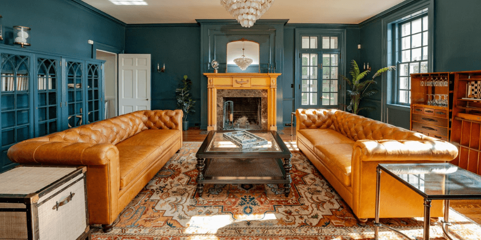 A wide view of Bohemia Manor Farm's teal library with two facing caramel Chesterfield sofas, a gold fireplace mantel with arched mirror, Gothic arch bookcases, capiz chandelier, Persian rug, and a wood bar cabinet by the windows.